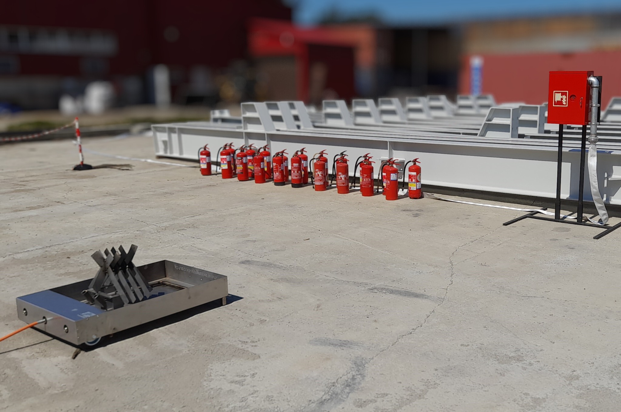 Stanowisko do ćwiczeń gaszenia pożarów z rzędem gaśnic i hydrantem w tle, na betonowej powierzchni. Sprzęt do symulacji ognia na pierwszym planie.