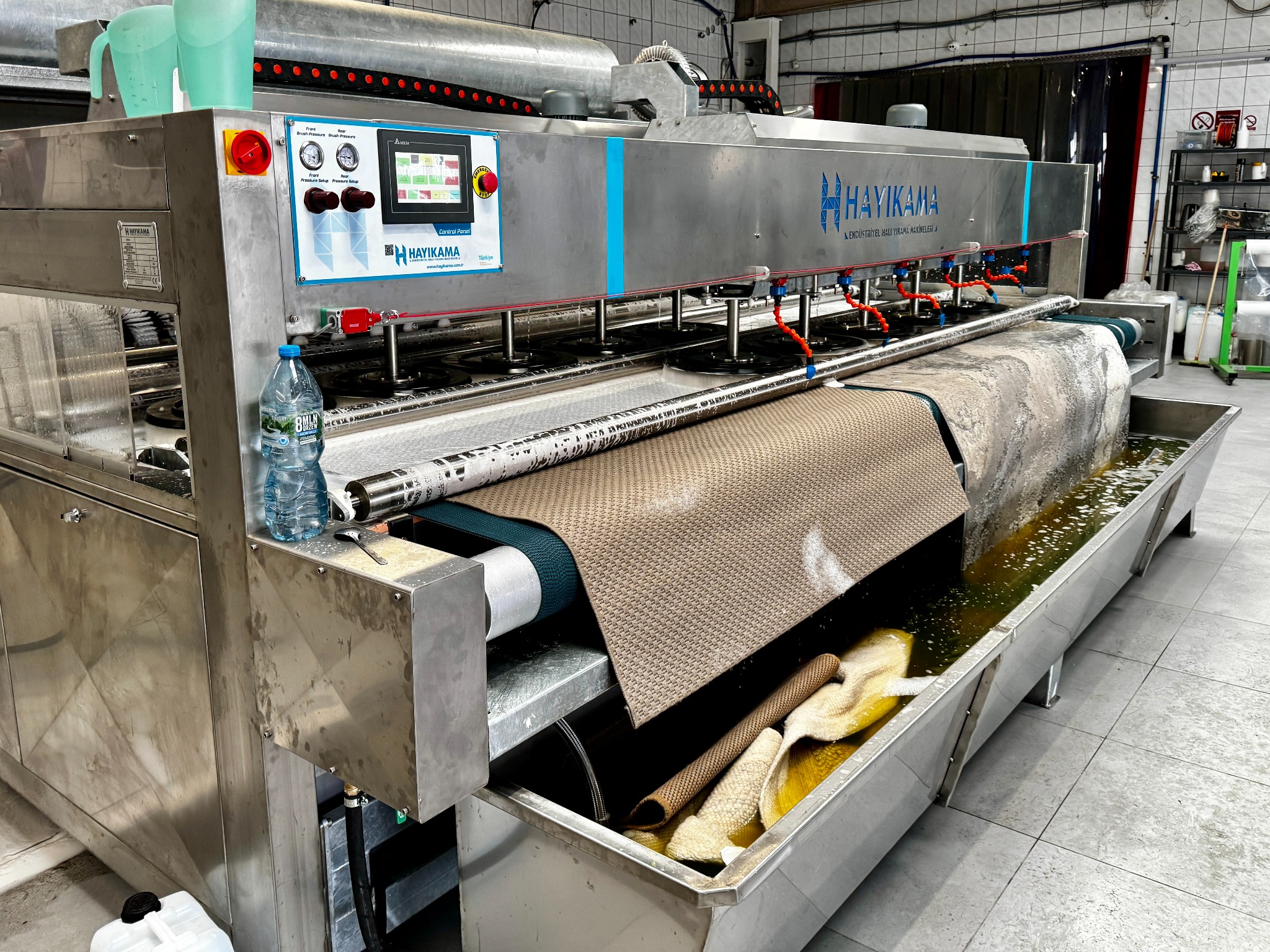 Profesjonalna maszyna HAYIKAMA do prania dywanów w trakcie pracy. Widoczne elementy sterujące i dywan przesuwający się przez urządzenie.