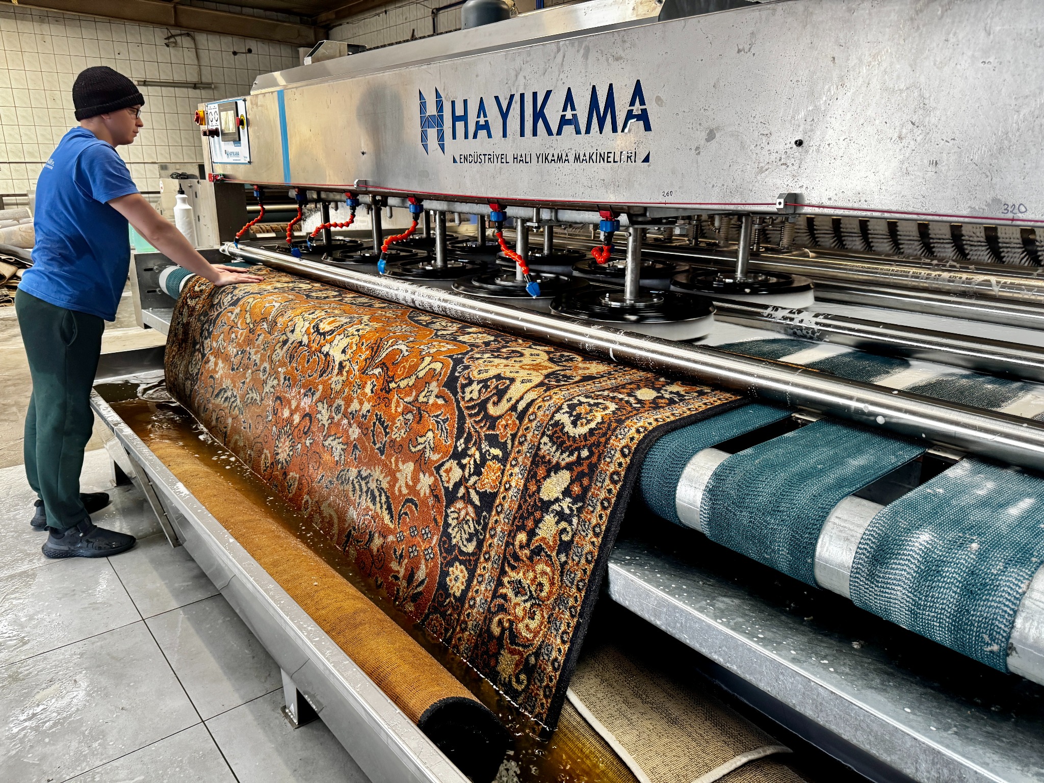 Pracownik obsługuje przemysłową maszynę do prania dywanów z wzorzystym, brązowo-pomarańczowym dywanem w trakcie czyszczenia. Widoczne dysze i szczotki maszyny.