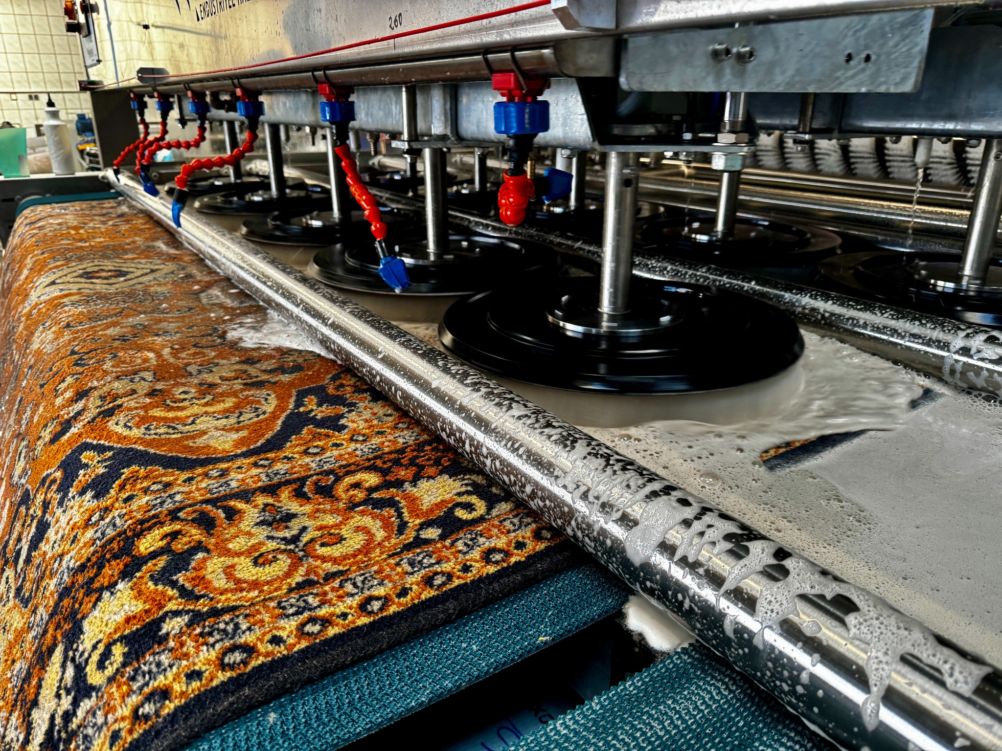 Automatyczna maszyna w trakcie prania wzorzystego dywanu. Widoczne szczotki, dysze i piana. Ujęcie z bliska, podkreślające detale i proces czyszczenia.