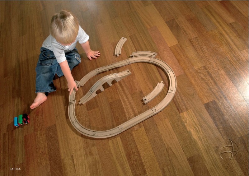 Chłopiec bawi się drewnianą kolejką na parkiecie Jatoba. Ciepłe odcienie drewna, naturalne światło. Widoczny fragment logo producenta w prawym dolnym rogu.