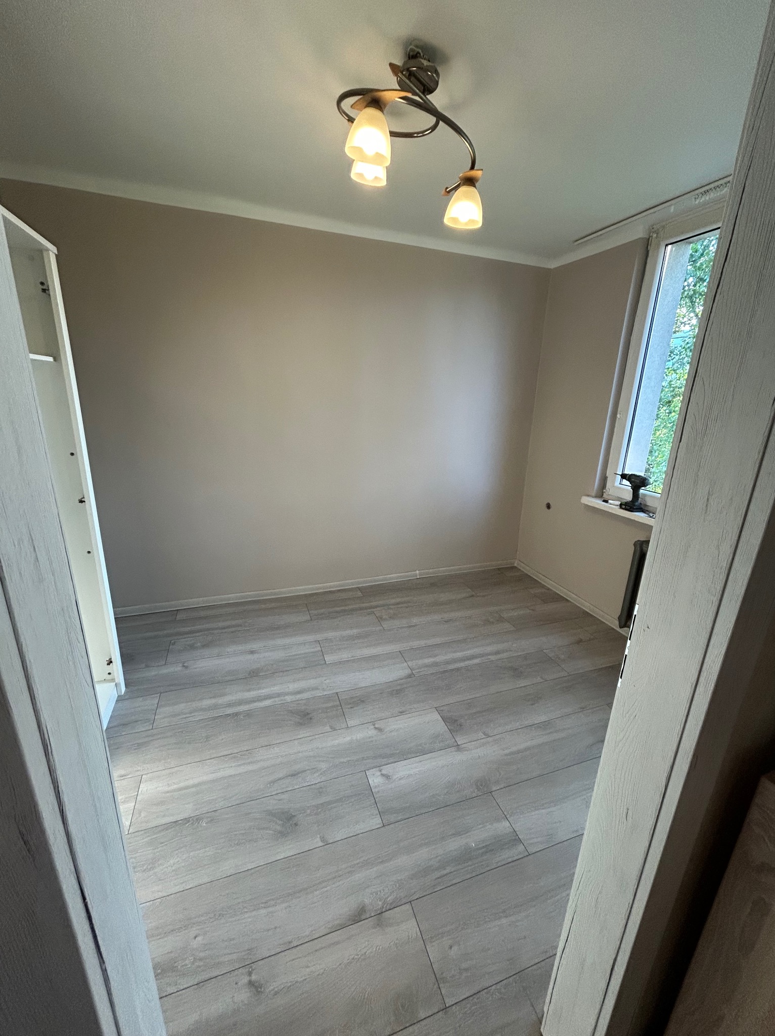 Jasne panele podłogowe ułożone w pokoju z beżowymi ścianami, widoczne przez framugę drzwi. Na parapecie leży wkrętarka.