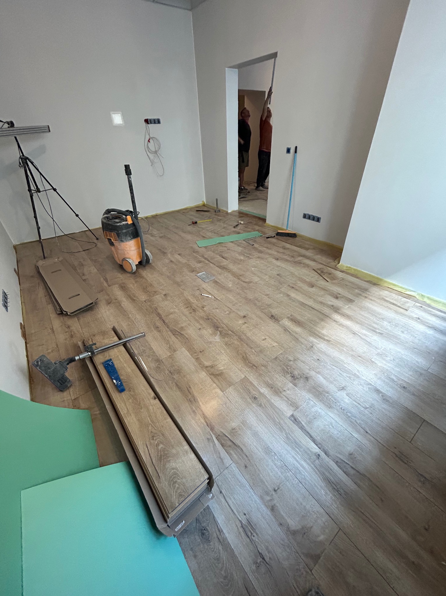 Wnętrze w trakcie montażu paneli podłogowych. Widoczne narzędzia, paczka paneli, odkurzacz przemysłowy i fragment ułożonej podłogi. W tle osoby pracujące przy ścianie.
