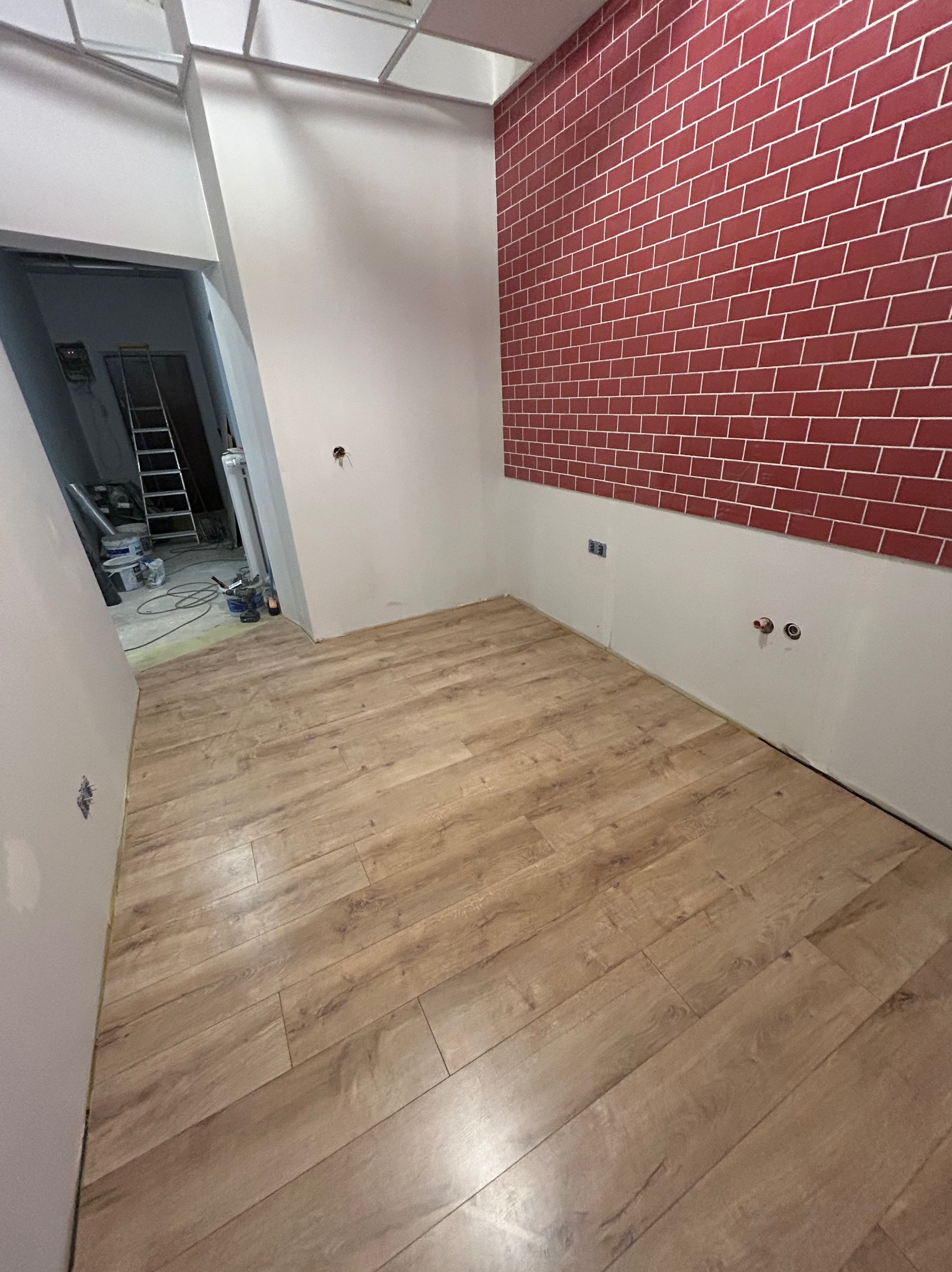 Jasne panele podłogowe ułożone w pokoju z czerwoną ścianą imitującą cegłę. W tle widoczne narzędzia i drabina, sugerujące trwający remont.
