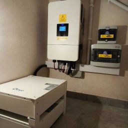 Te-Ka Energy Tomasz Strzyż - Biała bateria magazynu energii Deye, falownik i zabezpieczenia elektryczne na ścianie w pomieszczeniu. Widoczne przewody i rury instalacyjne.