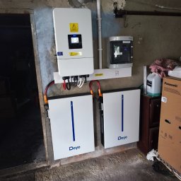 Te-Ka Energy Tomasz Strzyż - Zestaw magazynu energii Deye z inwerterem na ścianie otynkowanej piwnicy. Widoczne kable i przewody. Obok szafka z rozdzielnicą i karton Panasonic.