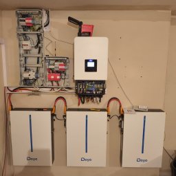 Te-Ka Energy Tomasz Strzyż - Instalacja magazynu energii Deye: trzy białe moduły bateryjne, inwerter i rozdzielnia elektryczna na beżowej ścianie, widoczne kable i narzędzia, co sugeruje etap montażu.