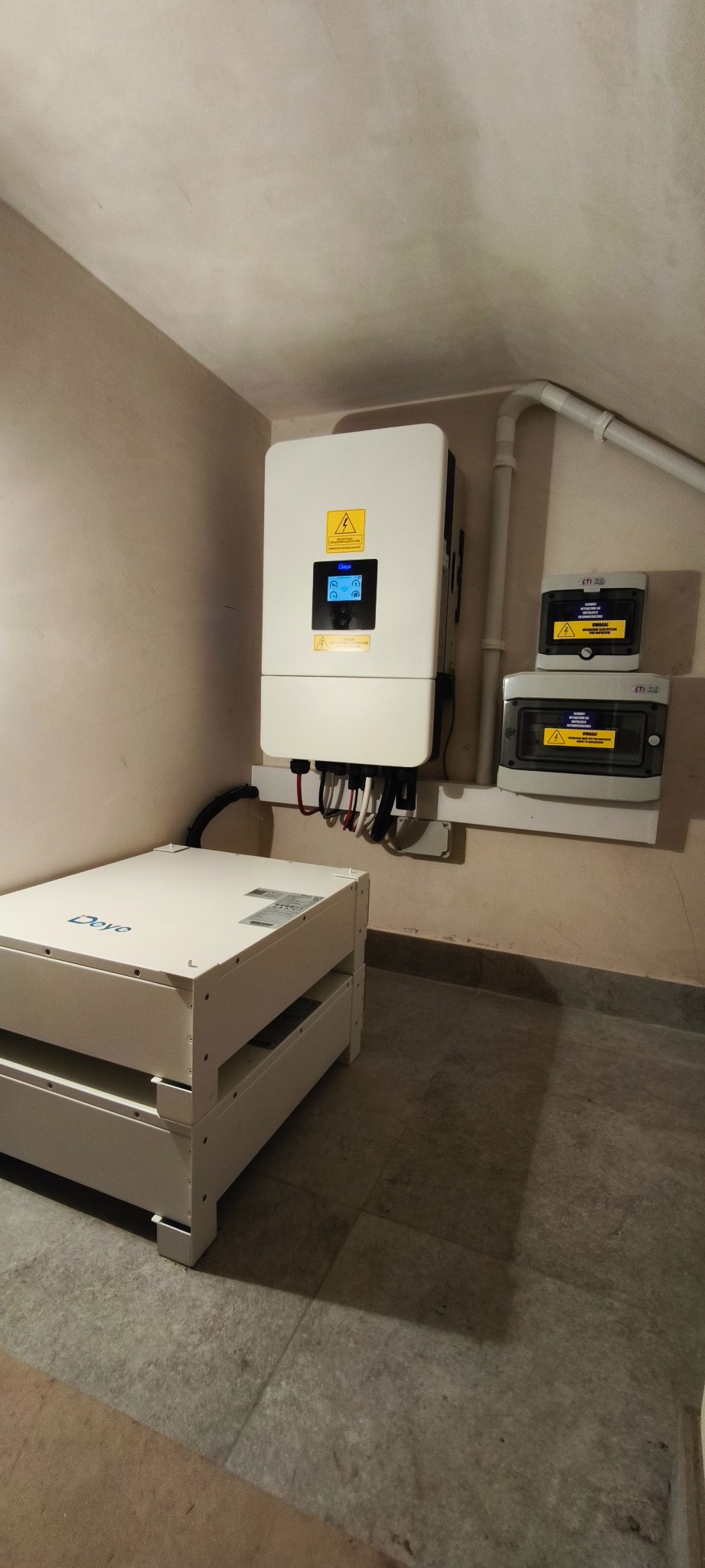 Biała bateria magazynu energii Deye, falownik i zabezpieczenia elektryczne na ścianie w pomieszczeniu. Widoczne przewody i rury instalacyjne.