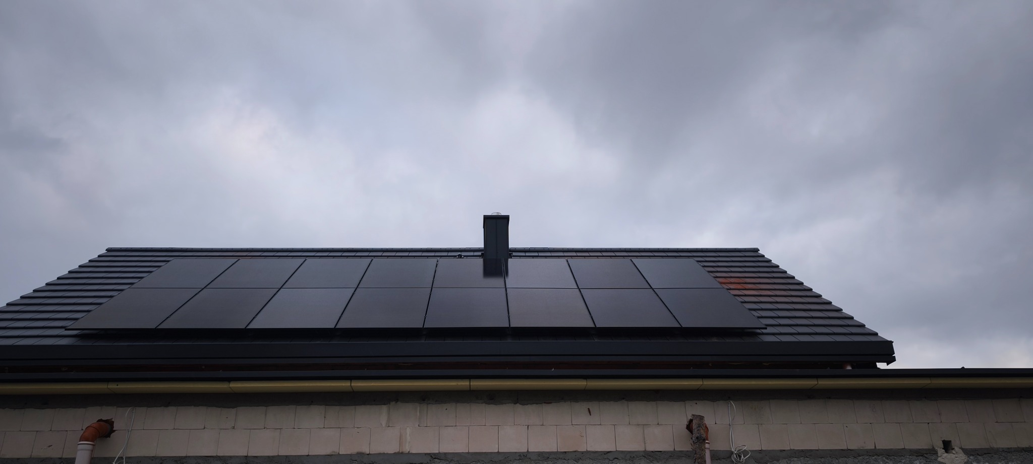 Panele fotowoltaiczne na dachu domu w Dojazdów, zamontowane na grafitowej dachówce. Widok na komin i pochmurne niebo.
