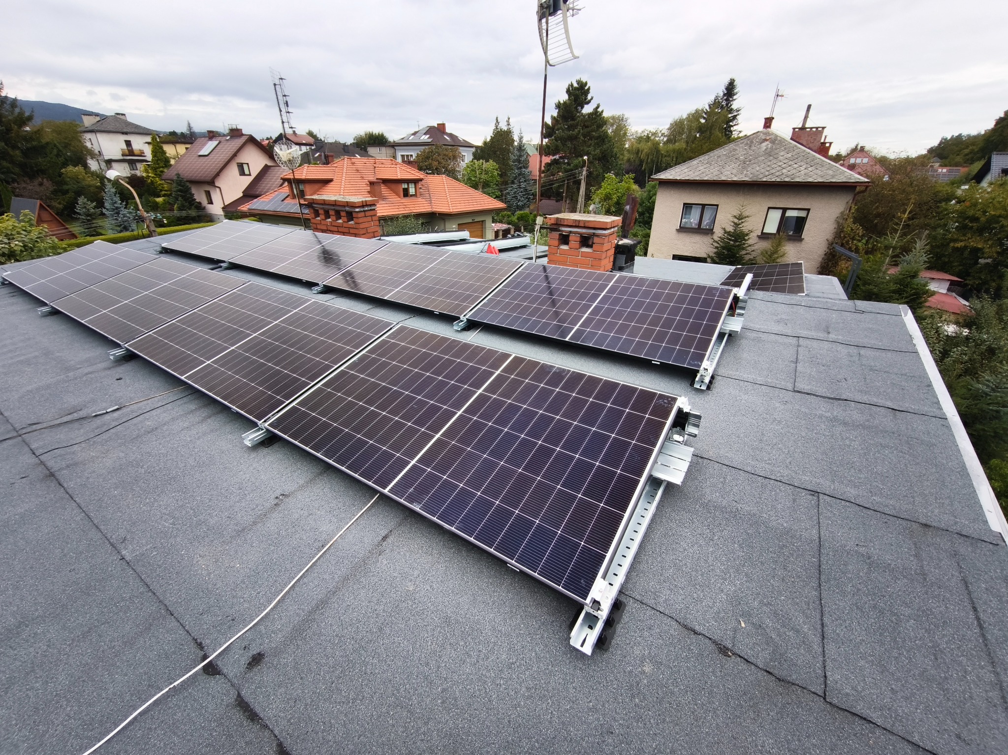 Panele fotowoltaiczne na dachu domu jednorodzinnego, widok z góry, instalacja w trakcie montażu, tło stanowią budynki mieszkalne i drzewa.