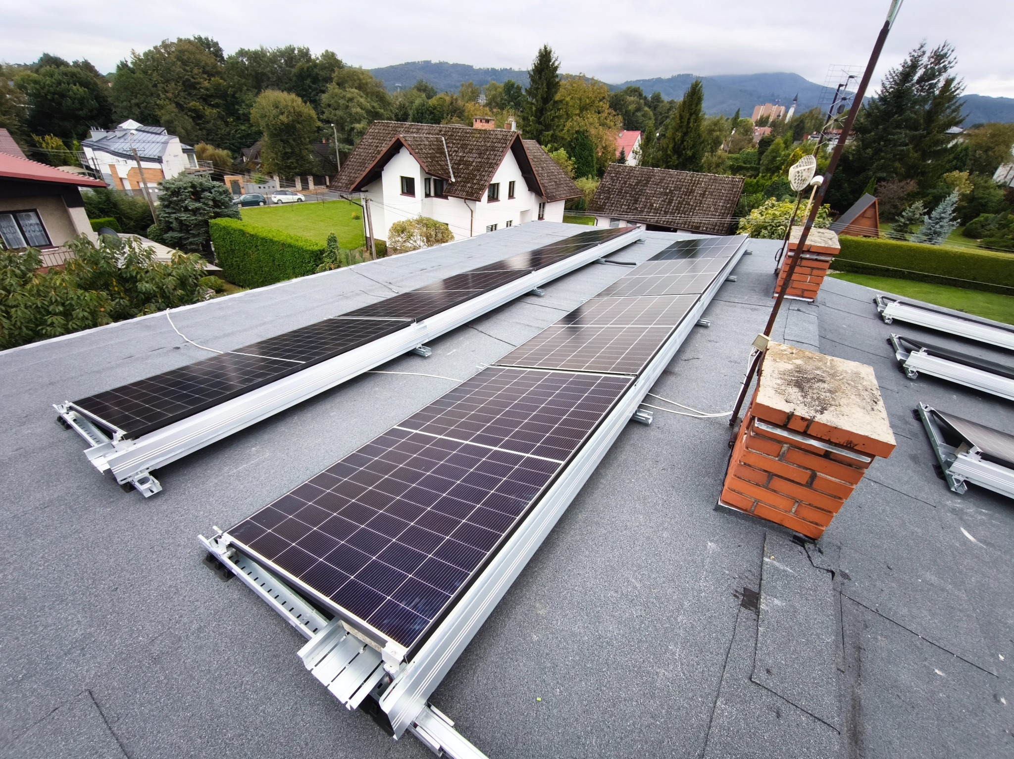 Panele fotowoltaiczne na dachu domu jednorodzinnego, widoczne kominy i antena, w tle zabudowa mieszkalna i zalesione wzgórza.