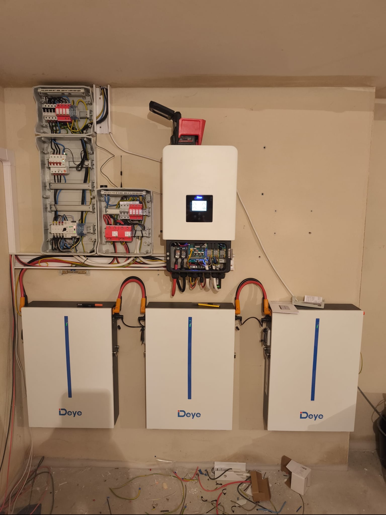 Instalacja magazynu energii Deye: trzy białe moduły bateryjne, inwerter i rozdzielnia elektryczna na beżowej ścianie, widoczne kable i narzędzia, co sugeruje etap montażu.