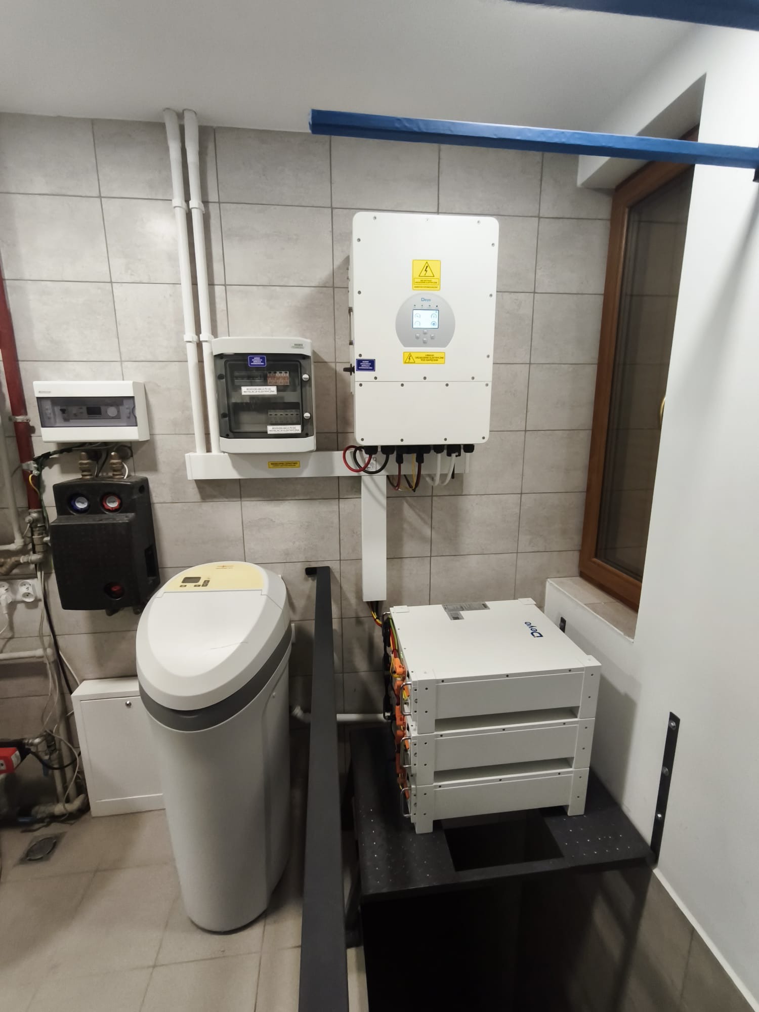 Kompletna instalacja fotowoltaiczna w pomieszczeniu: inwerter, magazyn energii Deye, rozdzielnice i zmiękczacz wody. Widok na szare płytki i okno.
