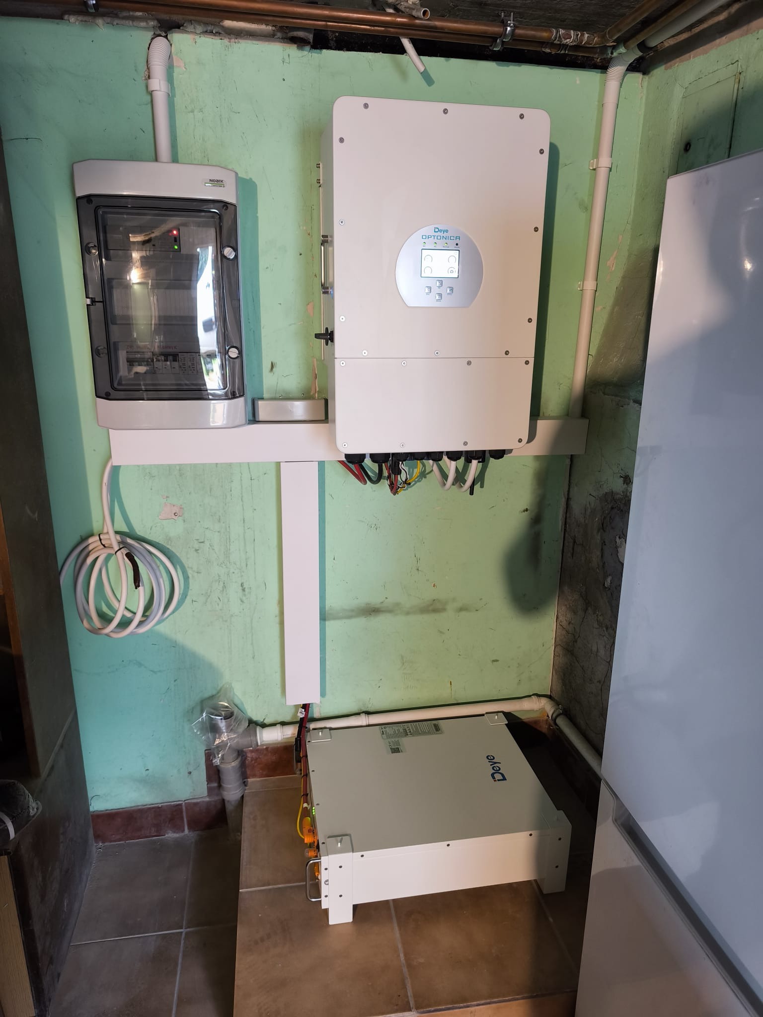 Zamontowany inwerter fotowoltaiczny Deye Optonica z rozdzielnicą na ścianie o zielonym odcieniu w piwnicy, widoczne kable i rury instalacyjne.