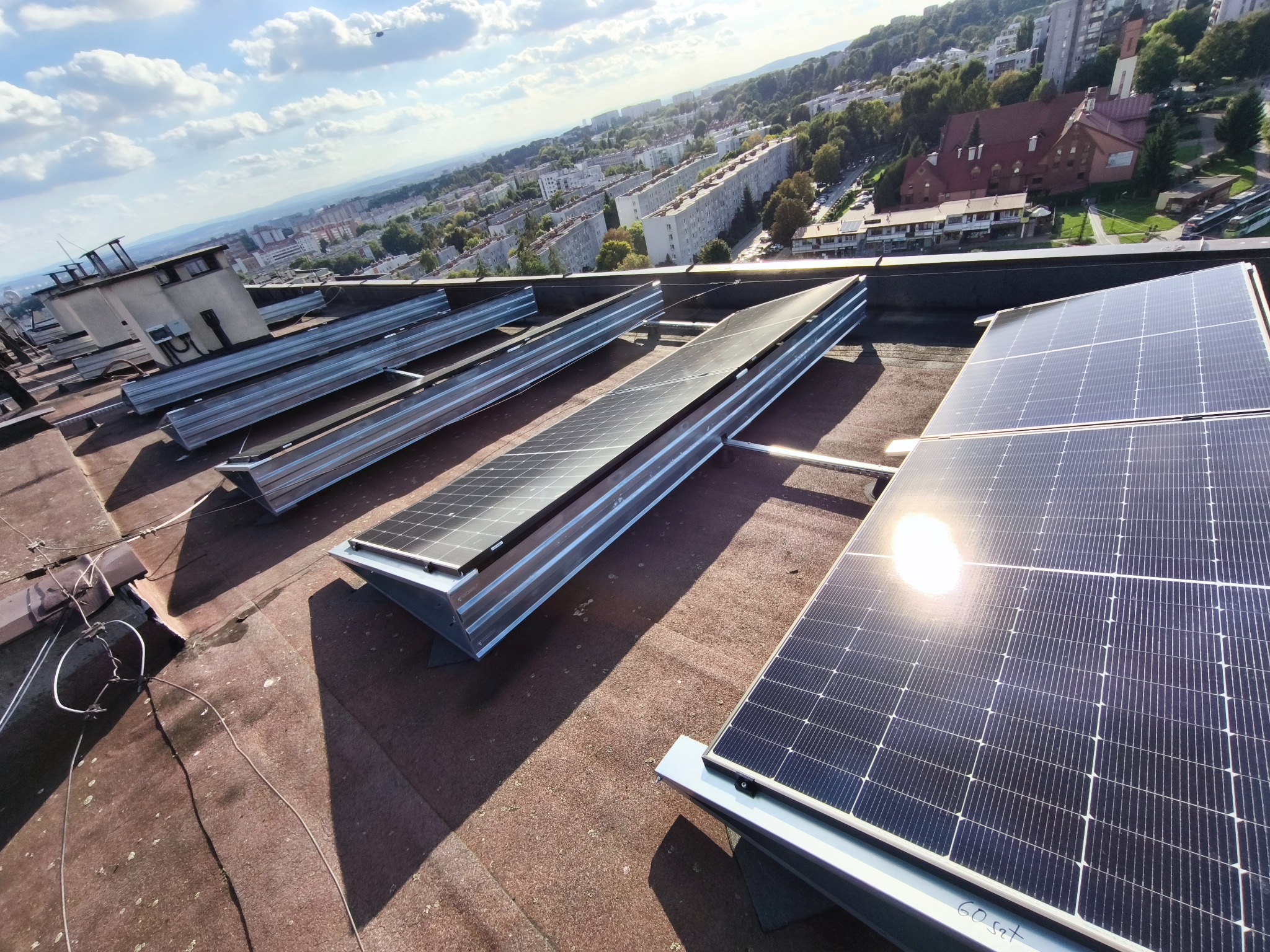 Montaż paneli słonecznych na dachu budynku z widokiem na miasto w tle. Widoczne metalowe konstrukcje wsporcze i częściowo zainstalowane panele. Słoneczny dzień.