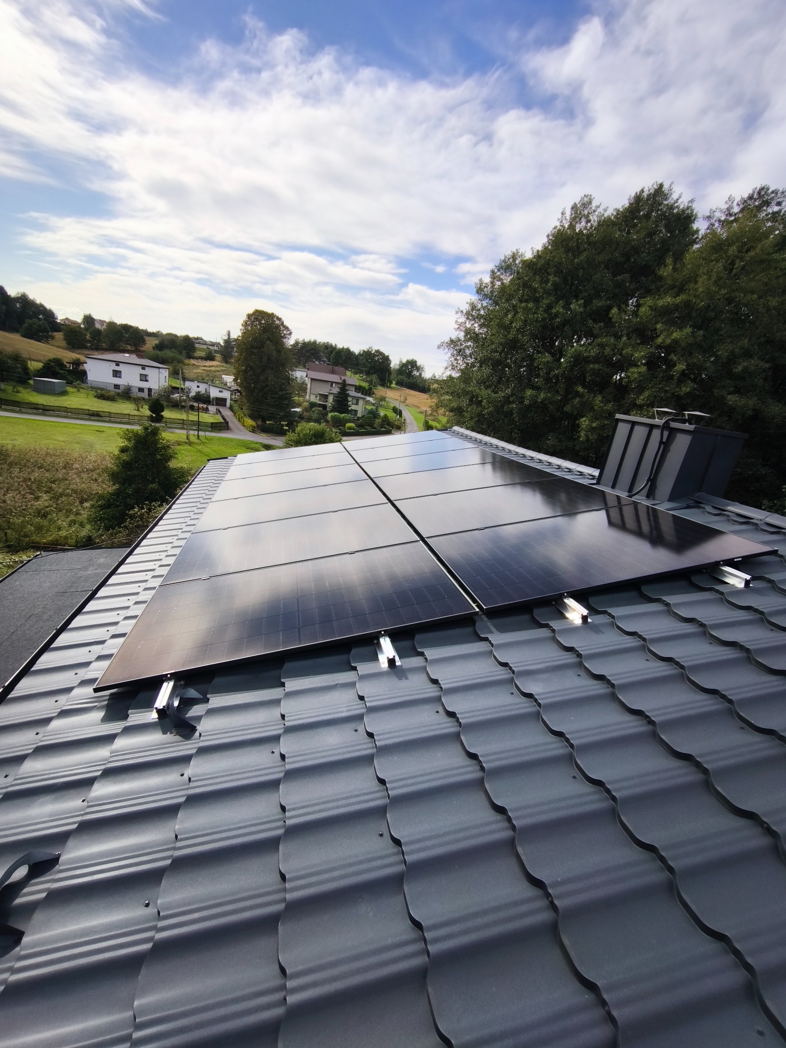 Cztery panele fotowoltaiczne na szarym dachu z blachodachówki, w tle domy i drzewa pod błękitnym niebem z chmurami. Widok z dołu, lekki kąt.