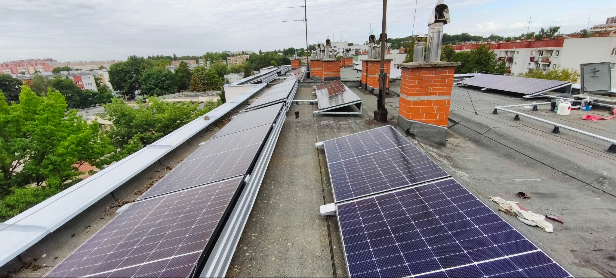Instalacja paneli słonecznych na dachu budynku z ceglanymi kominami w tle. Widok z góry, pochmurne niebo. Panele ułożone rzędami, częściowo pod kątem.