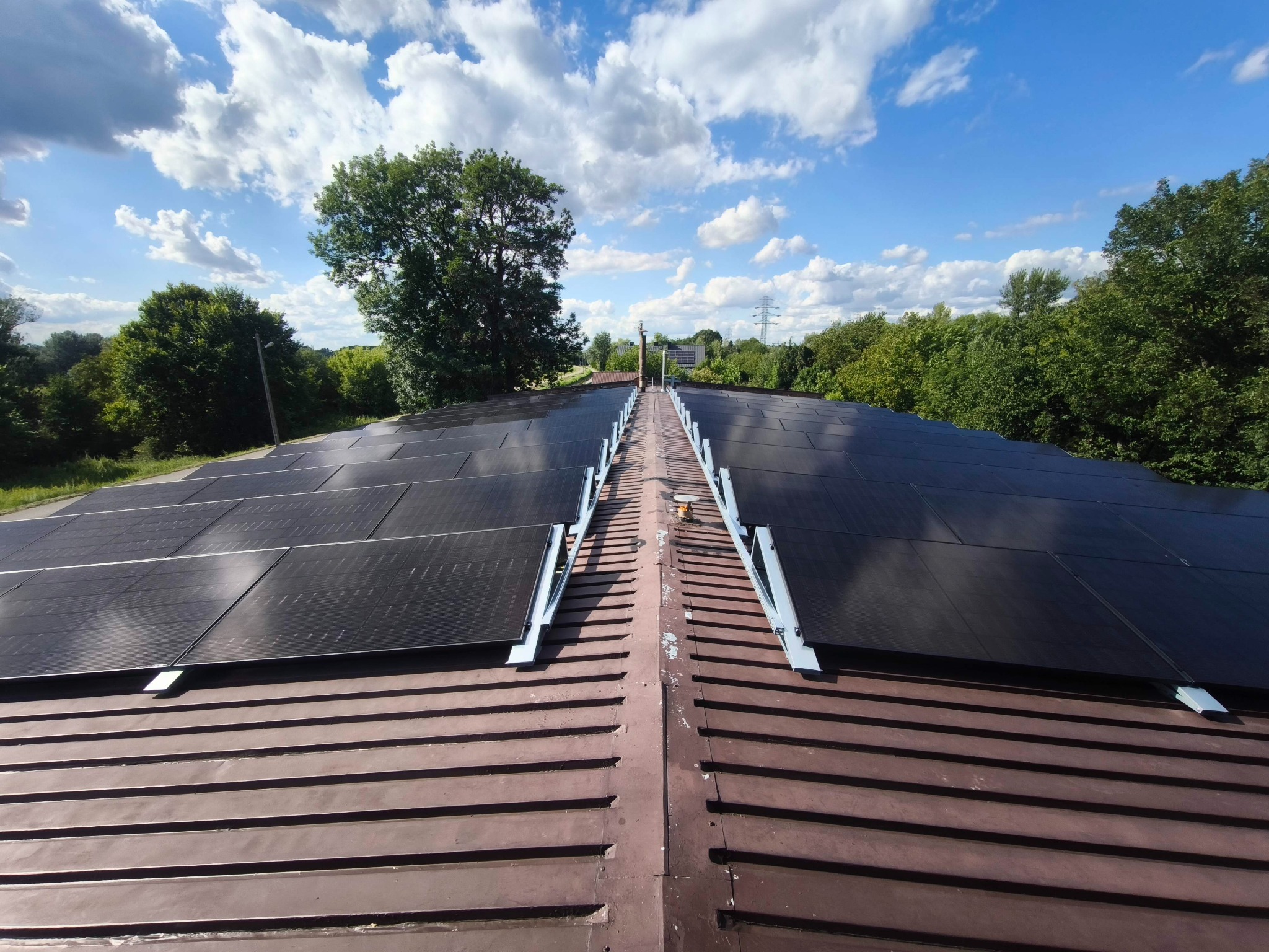 Widok z góry na brązowy dach z zamontowanymi panelami fotowoltaicznymi w słoneczny dzień. W tle drzewa i błękitne niebo z chmurami.