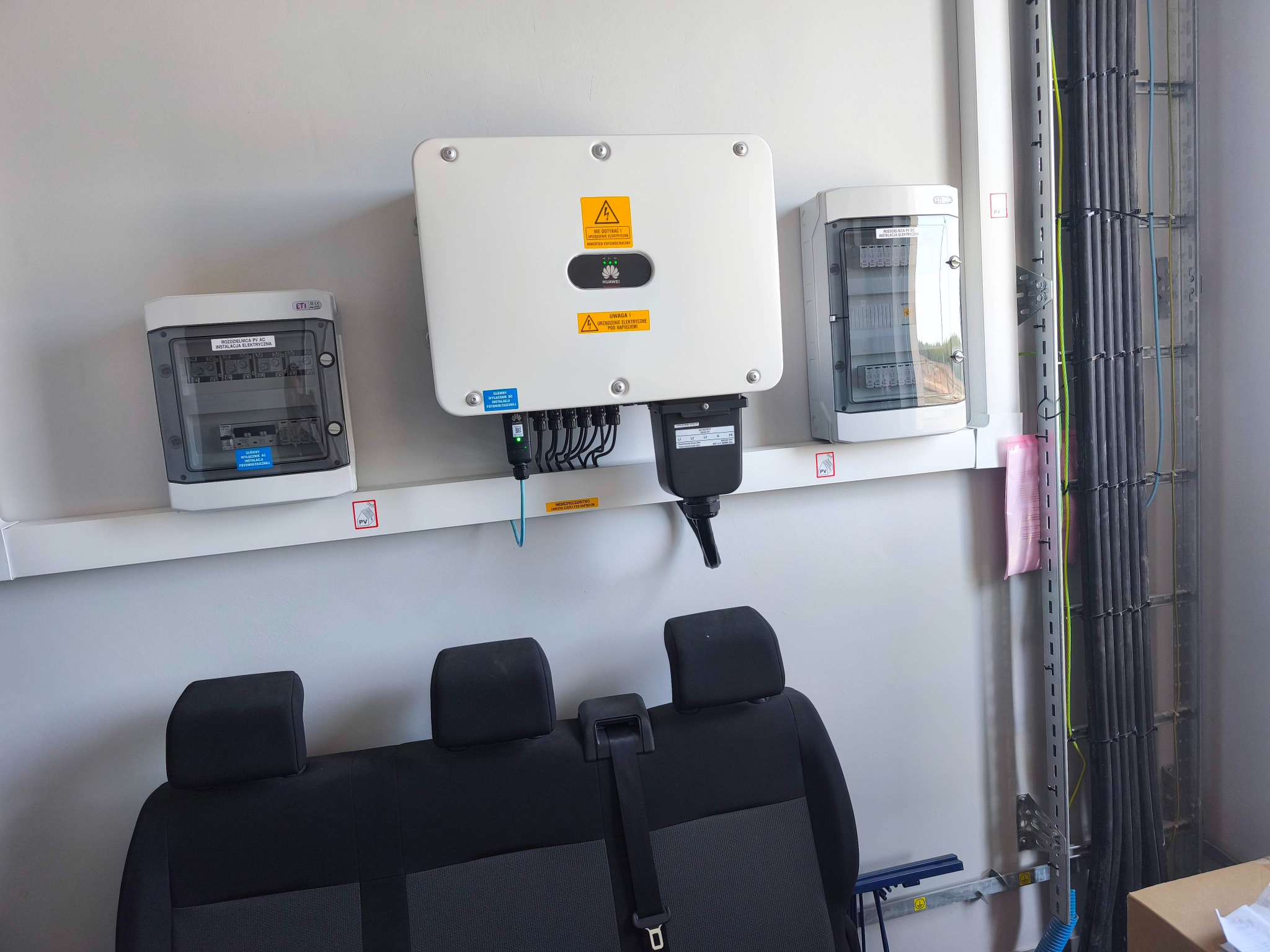 Biały inwerter fotowoltaiczny Huawei zamontowany na ścianie obok rozdzielnic PV AC. Widoczne kable i przewody. Na dole fragment czarnej kanapy samochodowej.