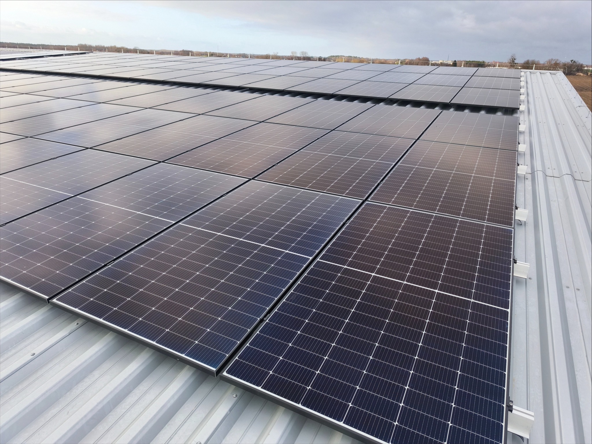 Rząd paneli słonecznych na metalowym dachu, widok z góry. Moduły fotowoltaiczne ułożone w regularny wzór, odbijające światło nieba. Widoczne elementy montażowe.