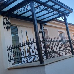 Sedzicki - Zadaszenie balkonu z metalową, dekoracyjną balustradą i lampami ściennymi. Konstrukcja w kolorze czarnym z przezroczystym dachem. Widok z dołu na elewację budynku.