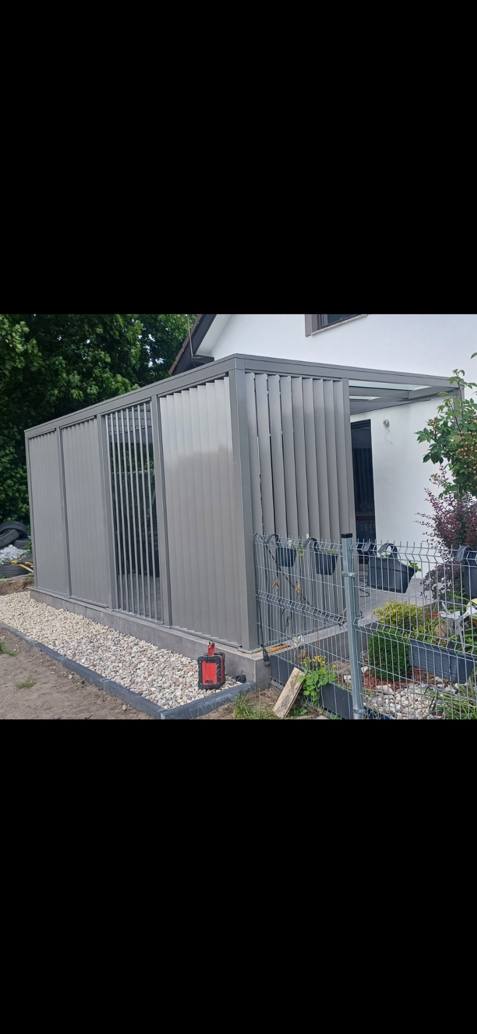 Szara pergola tarasowa ze stalową konstrukcją, ocynkowana ogniowo i malowana proszkowo, z żaluzjowymi panelami bocznymi, na tle białego domu i zieleni.