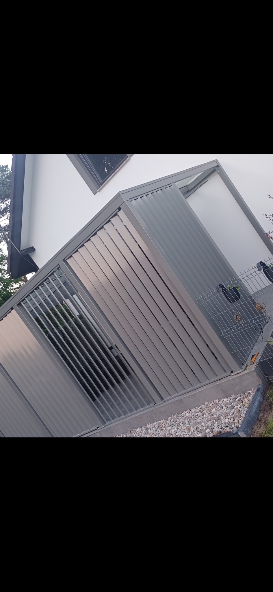 Szara pergola metalowa z lamelami, przylegająca do białego budynku. Widoczny fragment ogrodzenia z siatki i kamienny chodnik. Ujęcie z dołu, podkreślające konstrukcję.