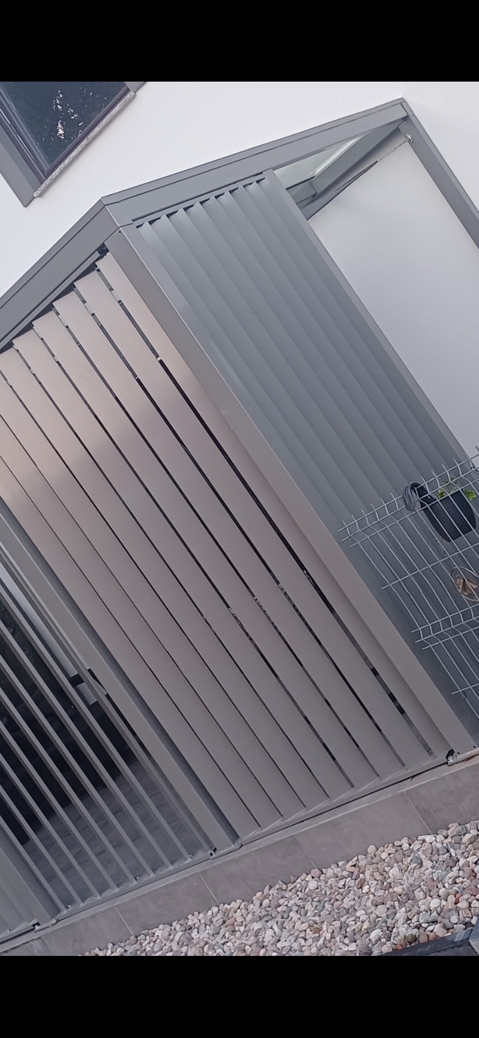 Szara, nowoczesna pergola z metalowymi lamelami, przymocowana do jasnej ściany budynku. Widoczne białe kamienie na podłożu i fragment doniczki.