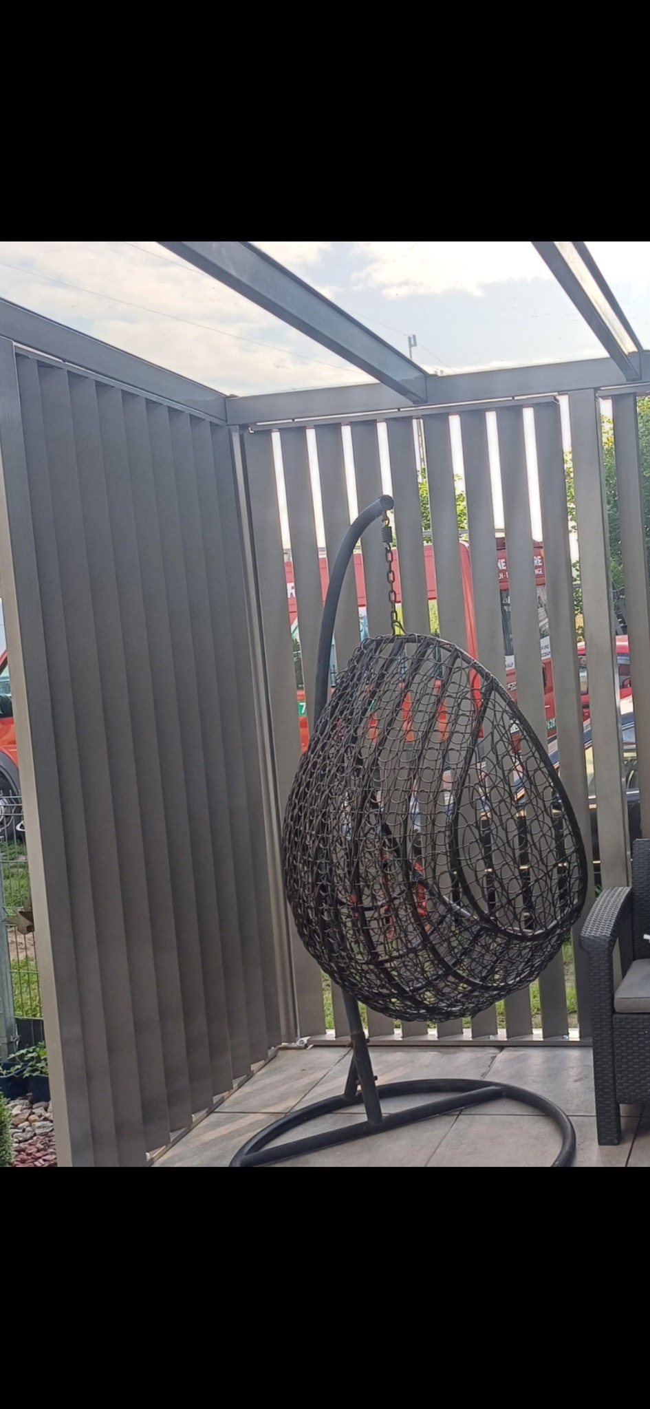 Zadaszona przestrzeń z metalowymi panelami i wiszącym fotelem typu kokon. Widoczne elementy konstrukcji z metalu. Fotel na tle pionowych paneli.