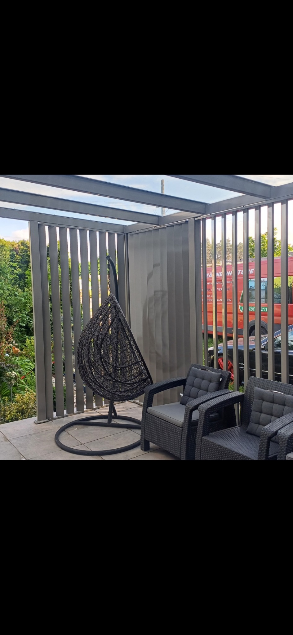 Zadaszona pergola z metalowymi, pionowymi lamelami tworząca zacienioną strefę relaksu z wiszącym fotelem i rattanowymi krzesłami.