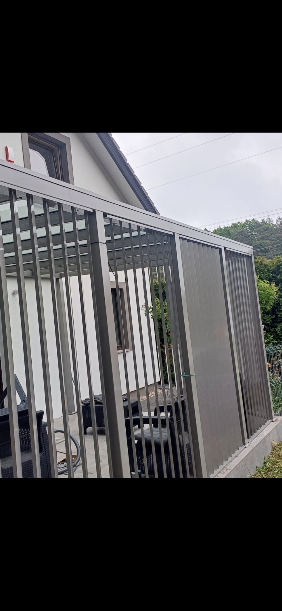 Nowoczesna pergola metalowa w kolorze szarym, z pionowymi panelami, tworząca zadaszone patio z widocznymi meblami ogrodowymi i fragmentem domu w tle.