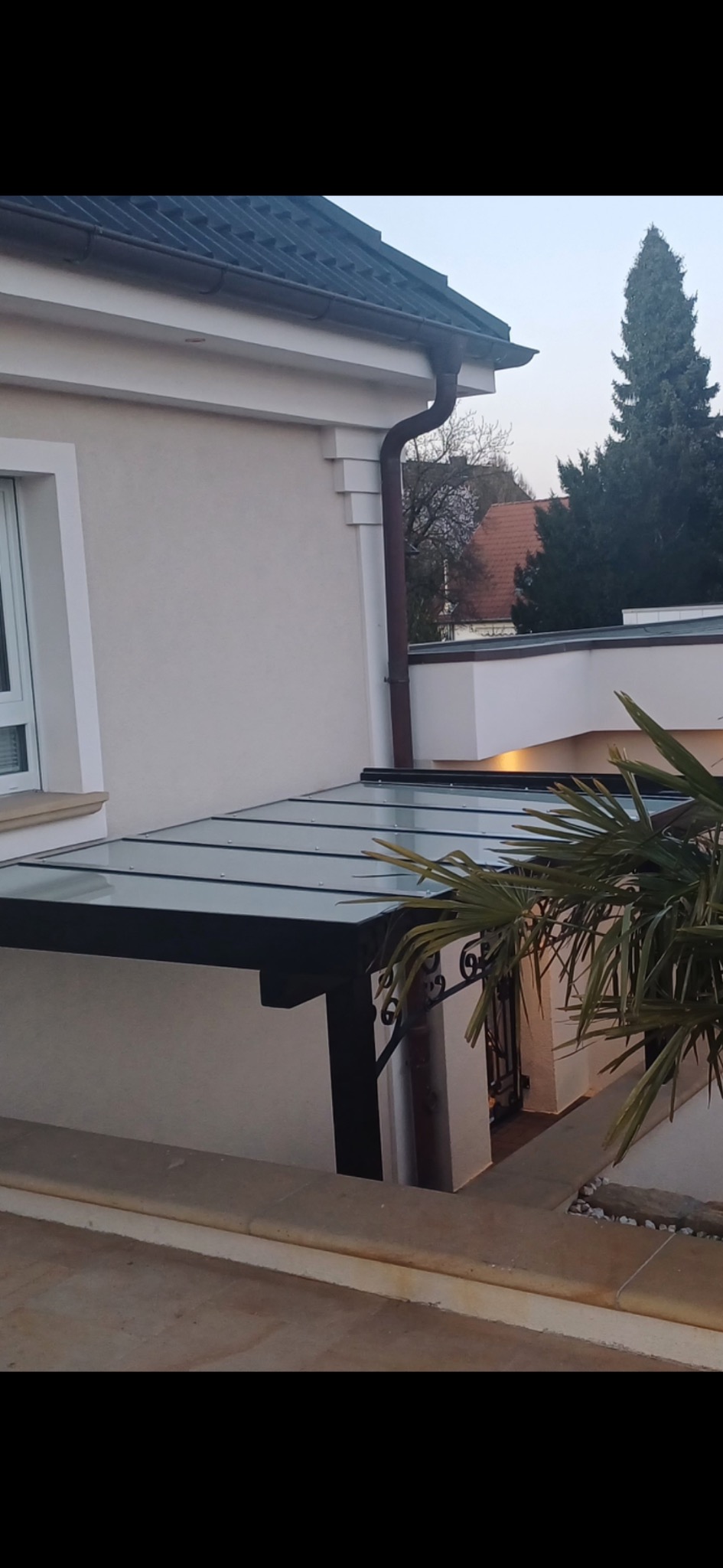 Czarna, metalowa pergola z przezroczystym dachem przylegająca do jasnej ściany budynku. Widoczne elementy dekoracyjne i fragment rośliny. Ujęcie z dołu.