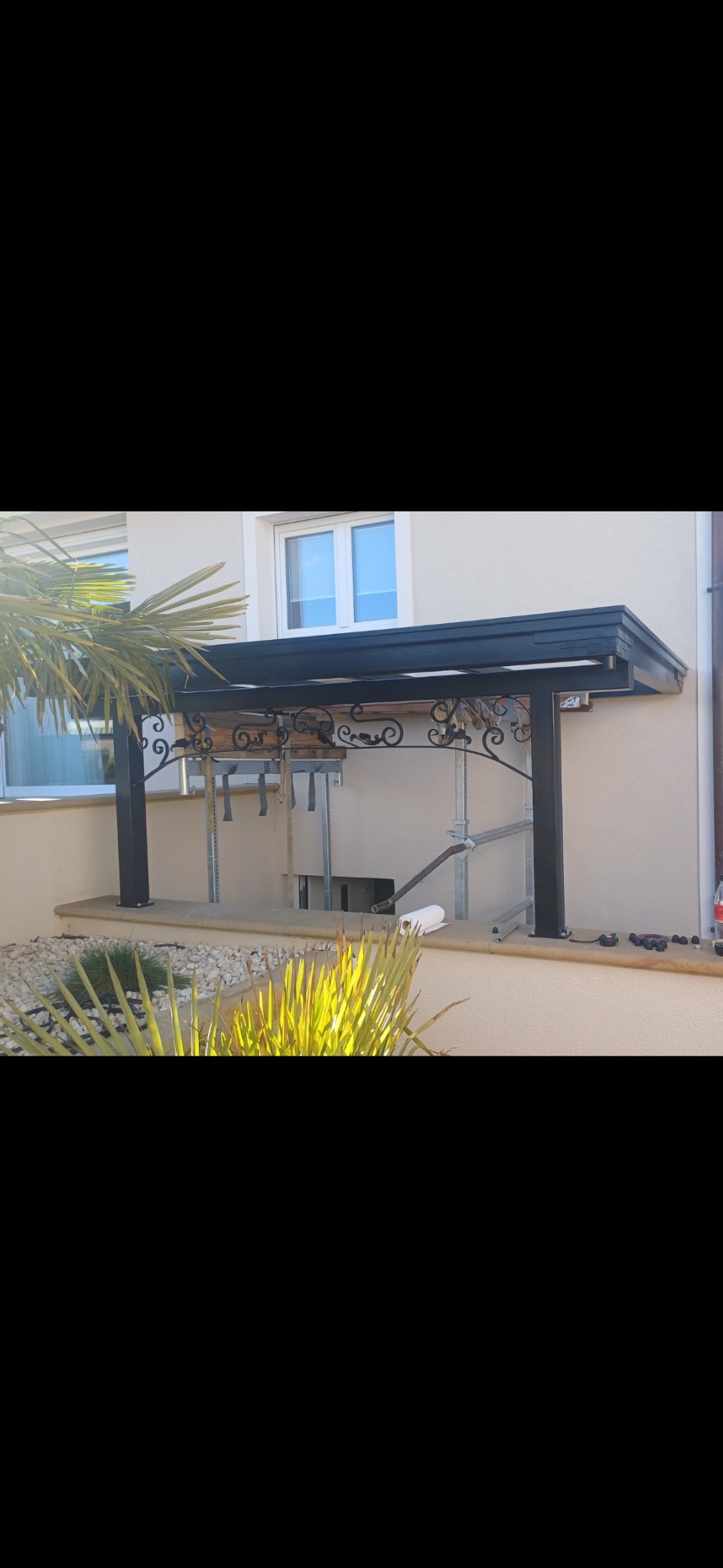 Czarna metalowa pergola z ozdobnymi elementami, przymocowana do ściany budynku. Widoczne rośliny w ogrodzie i fragment okna. Elementy montażowe pod konstrukcją.