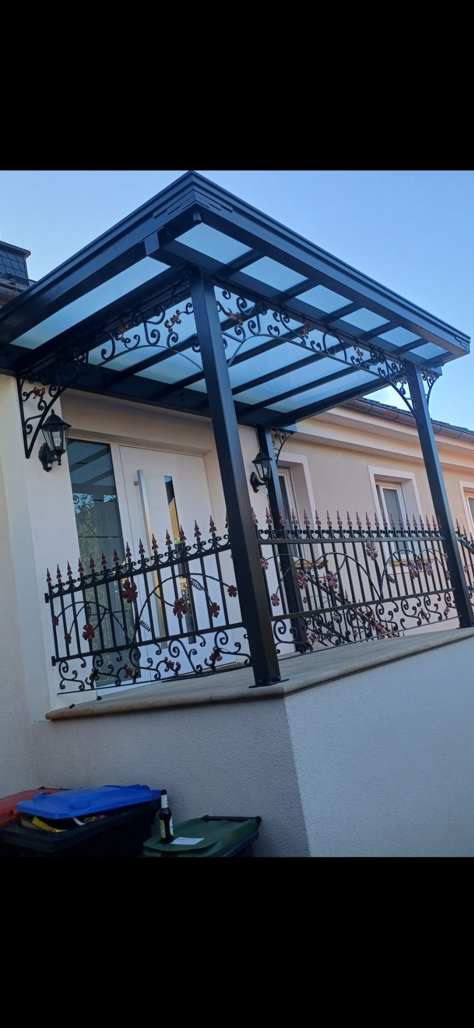 Zadaszenie balkonu z metalową, dekoracyjną balustradą i lampami ściennymi. Konstrukcja w kolorze czarnym z przezroczystym dachem. Widok z dołu na elewację budynku.