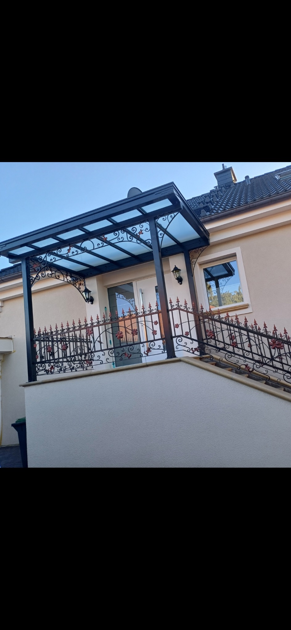 Czarna metalowa pergola z przezroczystym zadaszeniem nad wejściem do domu i balustrada z motywem róż na schodach. Eleganckie wykończenie.