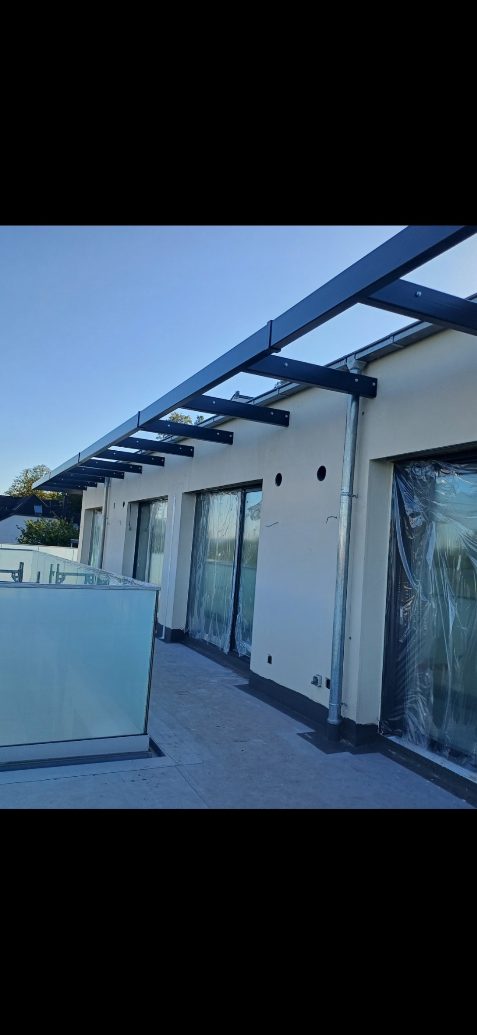 Czarna pergola metalowa przymocowana do ściany budynku z przeszkloną balustradą. Okna zabezpieczone folią ochronną.