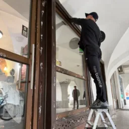 Mężczyzna na drabinie czyści okno witryny sklepowej z resztek farby, widoczne plamy zabrudzeń na brązowej ramie okiennej i szybie, na zewnątrz arkady.