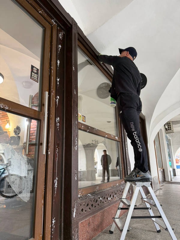 Mężczyzna na drabinie czyści okno witryny sklepowej z resztek farby, widoczne plamy zabrudzeń na brązowej ramie okiennej i szybie, na zewnątrz arkady.