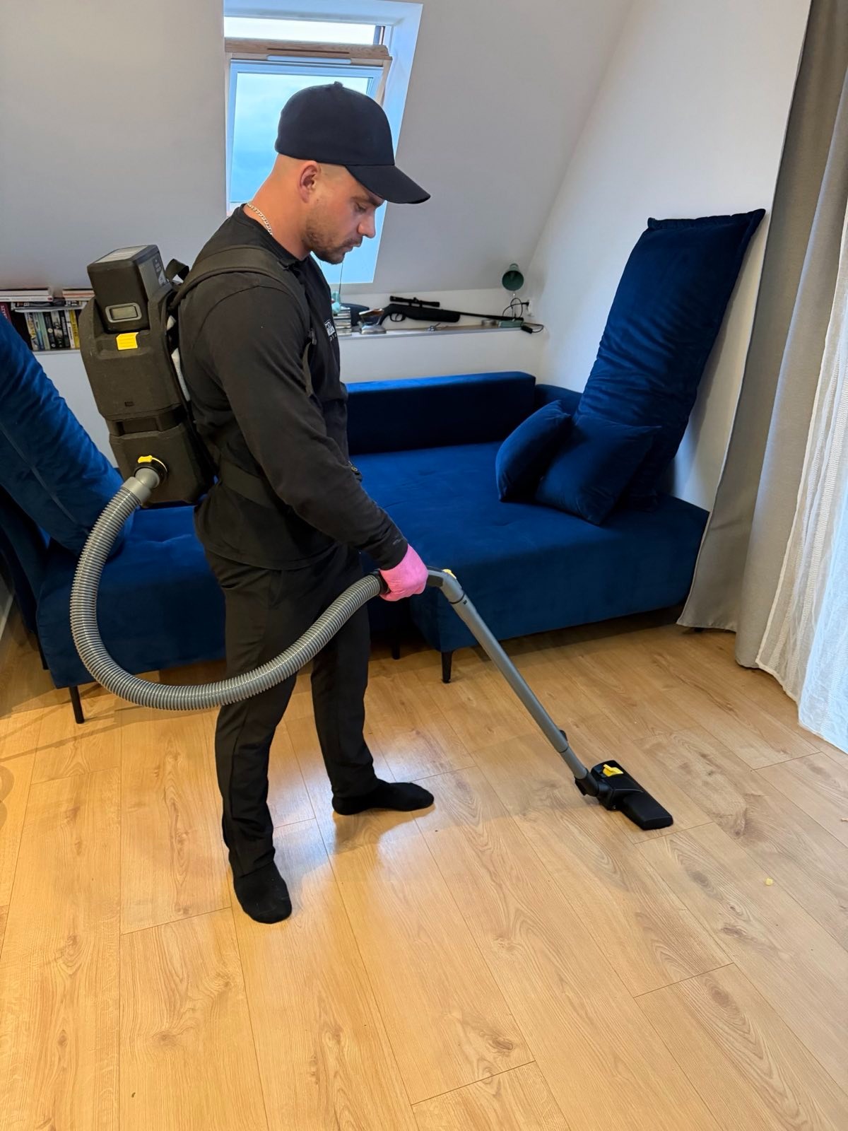 Mężczyzna w czarnym stroju i różowych rękawicach odkurza drewnianą podłogę za pomocą odkurzacza plecakowego w pokoju z niebieską sofą i oknem na poddaszu.