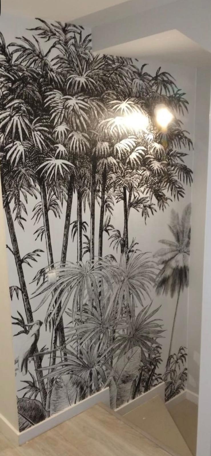 Ściana pokryta tapetą z motywem tropikalnych drzew i ptaka, utrzymana w czarno-białej kolorystyce, oświetlona od góry, widoczny fragment schodów.