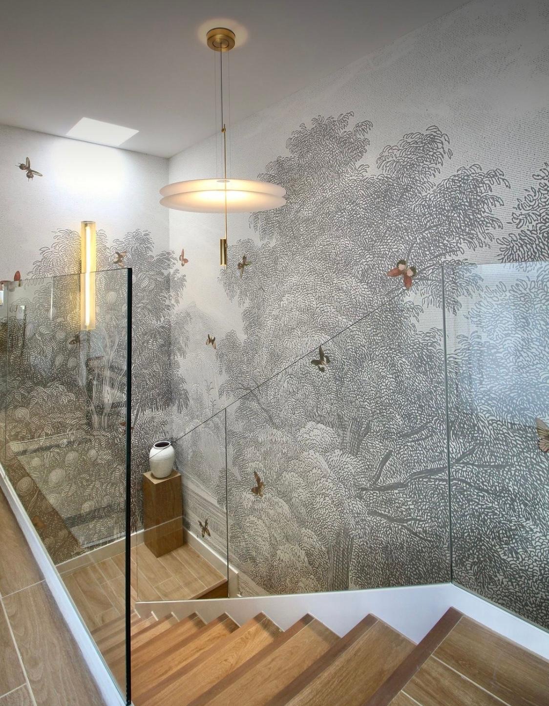 Klatka schodowa z fototapetą przedstawiającą leśny pejzaż, szklane balustrady i drewniane schody. Oświetlenie punktowe i wisząca lampa dodają elegancji wnętrzu.