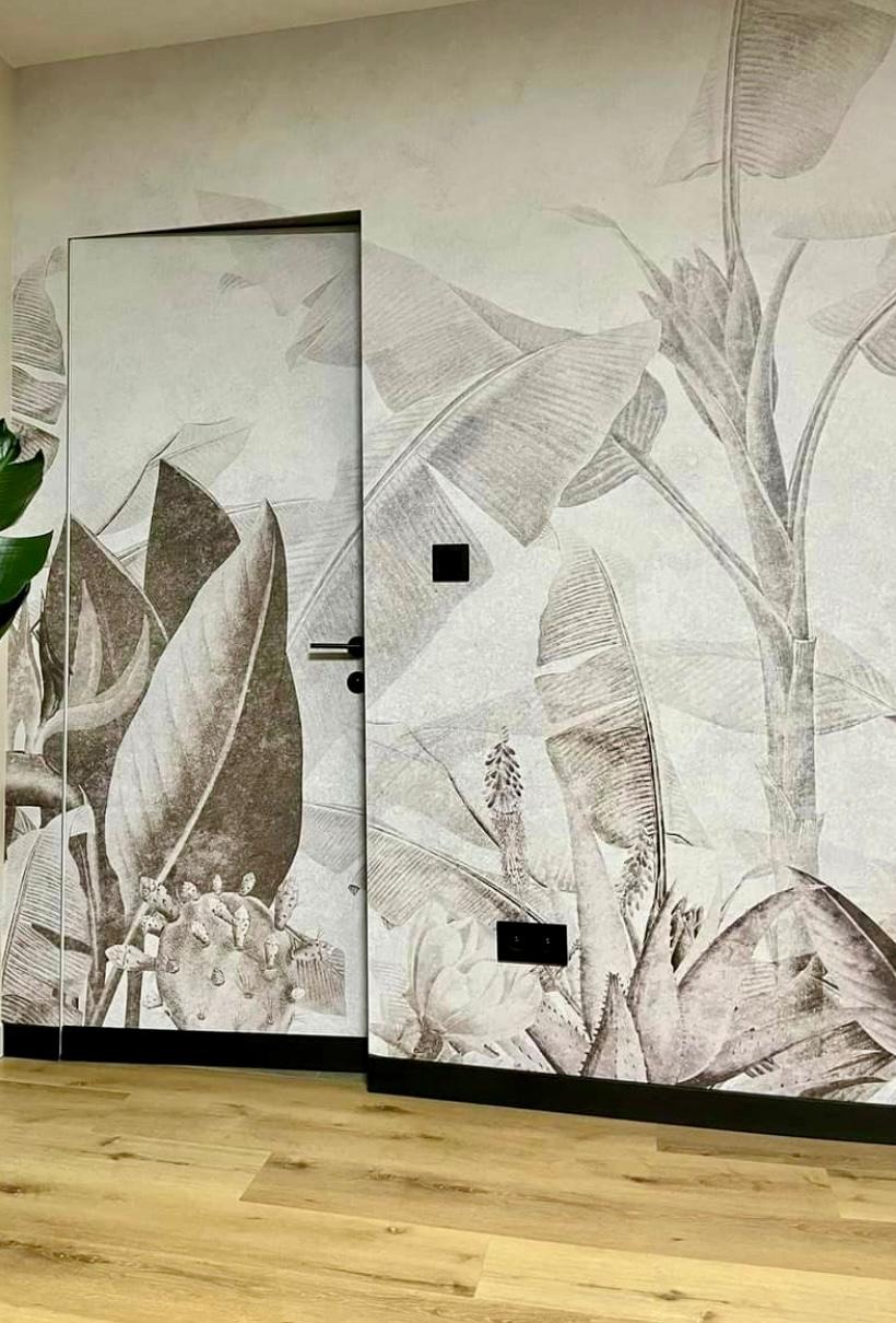 Tapeta z motywem tropikalnych liści na ścianie i drzwiach, z widocznymi włącznikami. Podłoga z paneli w ciepłym odcieniu drewna. Styl botaniczny, monochromatyczny.
