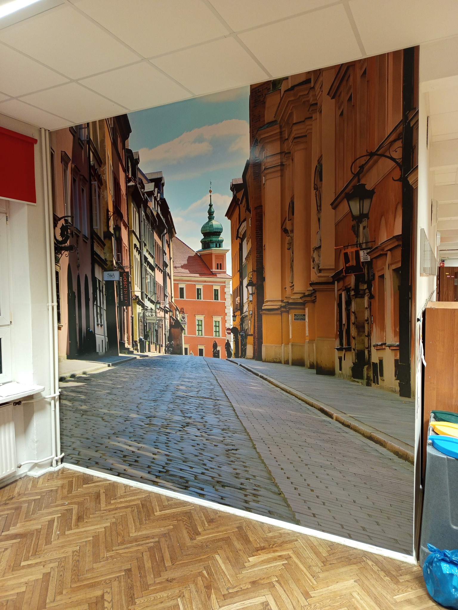 Fototapeta z motywem Starego Miasta w Warszawie, naklejona na ścianę w pomieszczeniu z parkietem w jodełkę. Widoczny fragment okna i grzejnika.