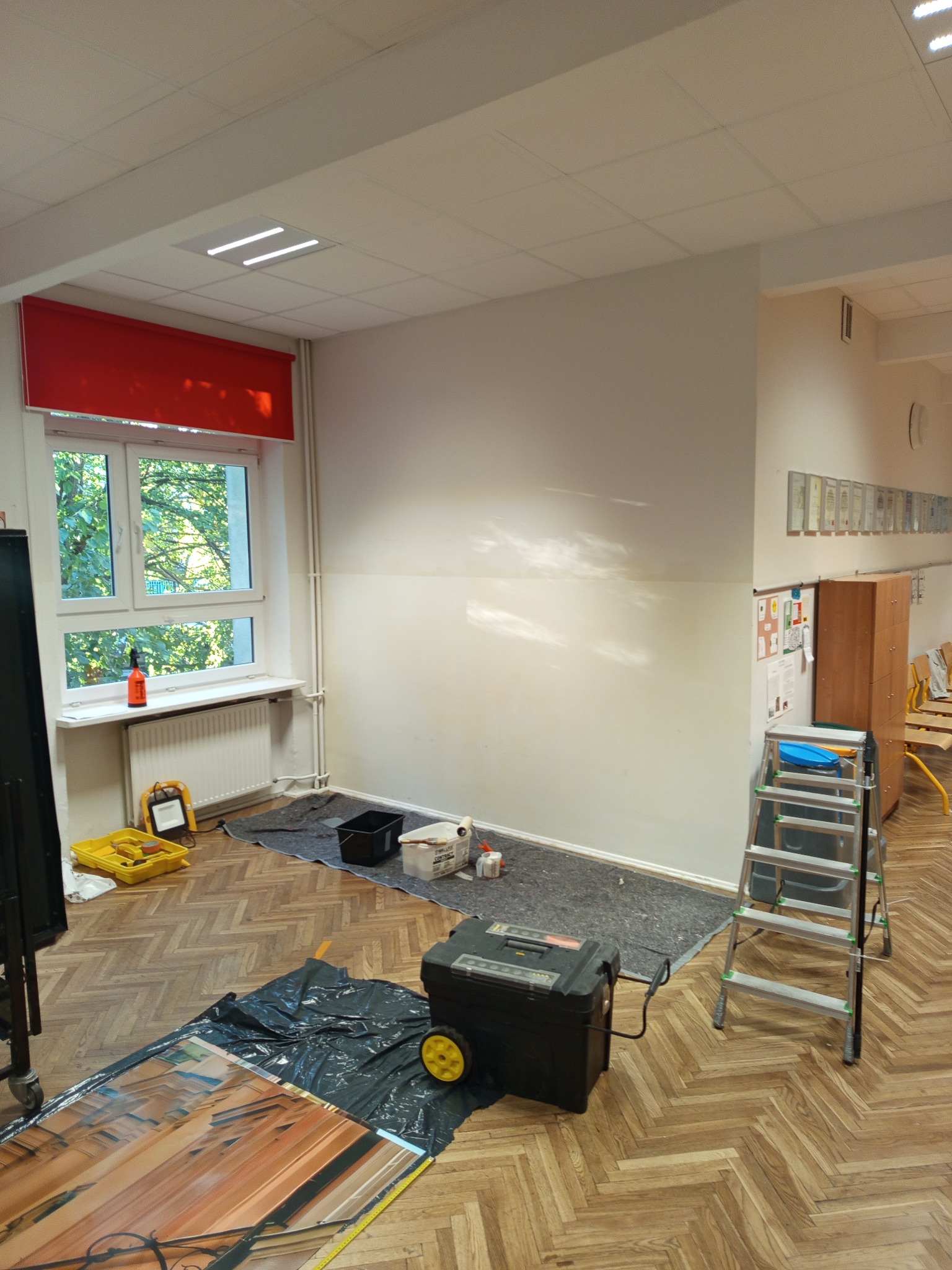 Przygotowania do malowania ścian w pomieszczeniu z parkietem. Widoczne narzędzia, drabina i zabezpieczona podłoga. Ściana częściowo przygotowana do prac.