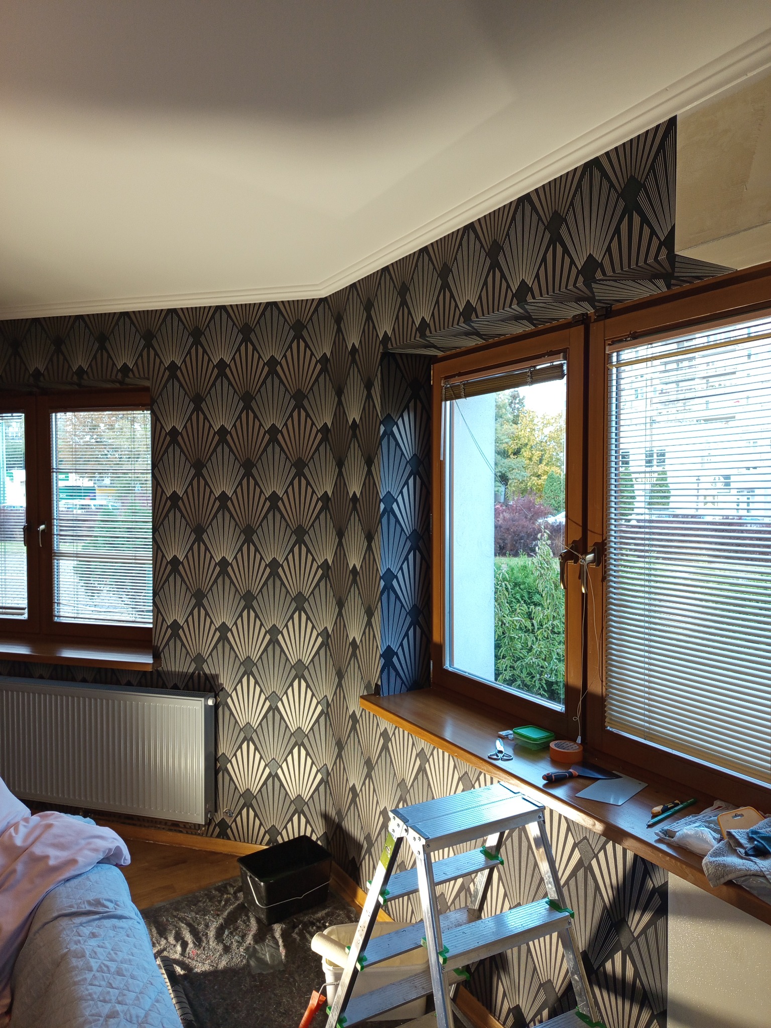 Art deco wzór na ścianie po remoncie. Widoczne okna z żaluzjami, drabina i narzędzia malarskie. Częściowo widoczny grzejnik i łóżko.