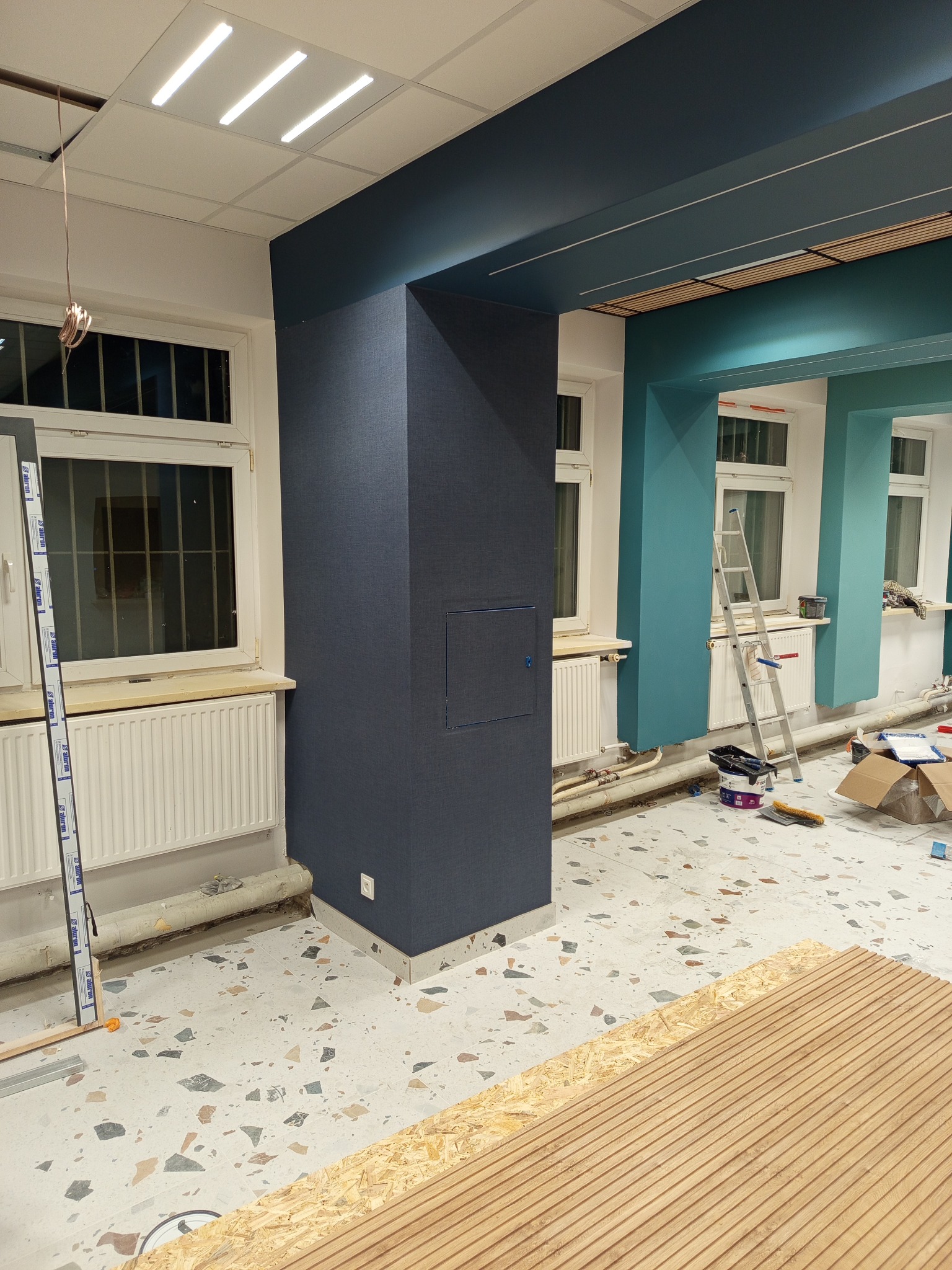 Wnętrze w trakcie prac wykończeniowych: kolumna obita tkaniną, pomalowany sufit, drabina, narzędzia malarskie i płyty na podłodze. Widok na odnowione pomieszczenie.