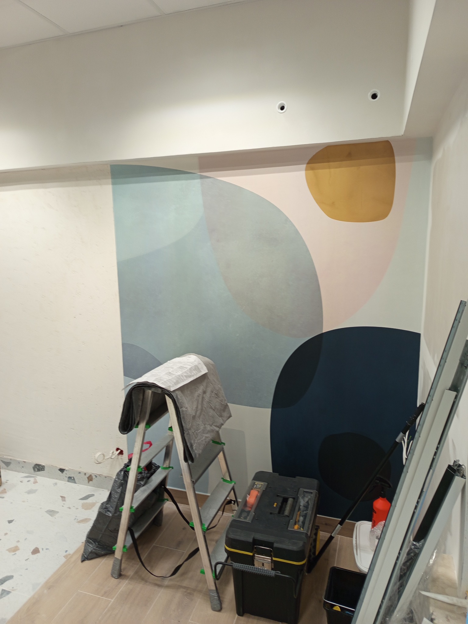 Tapetowanie ściany z abstrakcyjnym wzorem. Widoczna drabina, narzędzia i fragment podłogi. Praca w toku, nowoczesny design, jasne wnętrze.