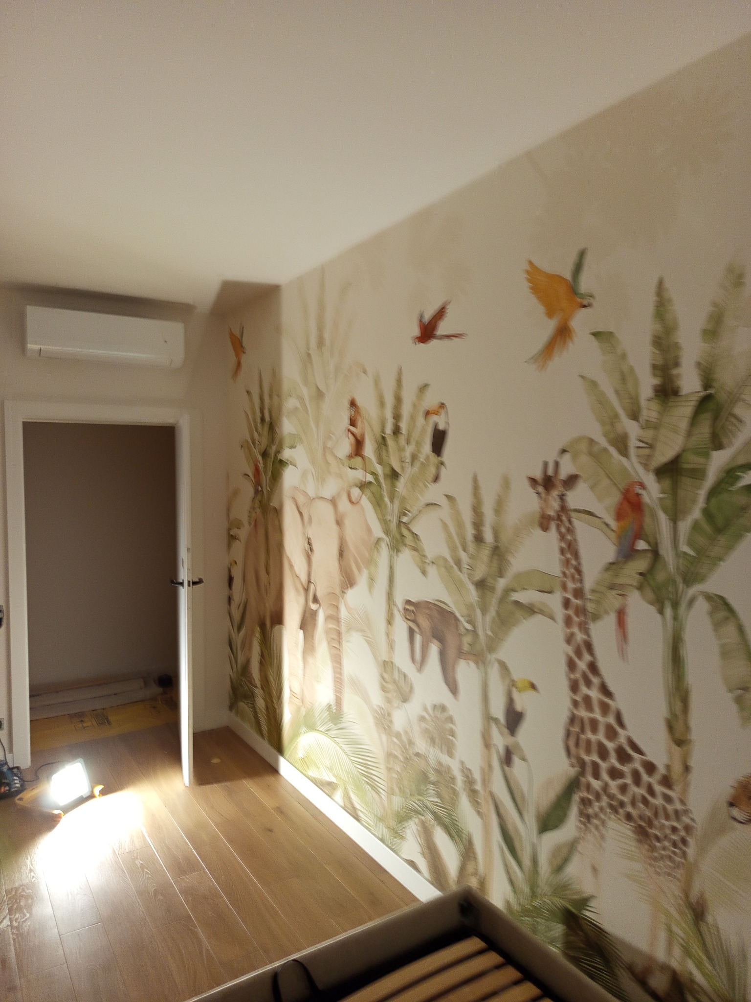 Pokój po dekoracji ścian: fototapeta z motywem dżungli z żyrafą, słoniem i papugami. Jasne panele na podłodze, widoczny fragment łóżka i lampa halogenowa.