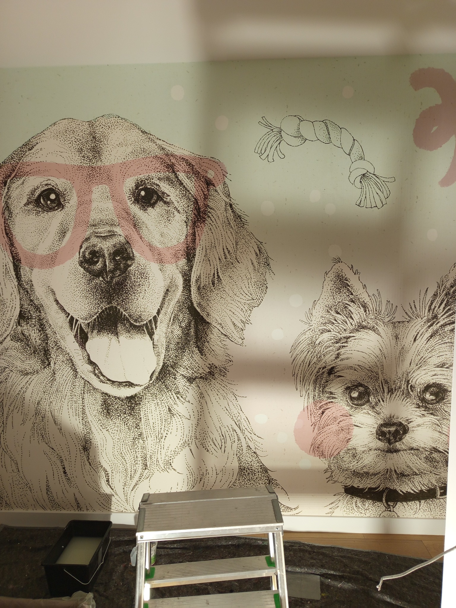 Ściana z tapetą w dziecięcym stylu: rysunki psów (golden retriever w okularach i york) oraz zabawki. Widoczny fragment drabiny i kuweta z klejem.
