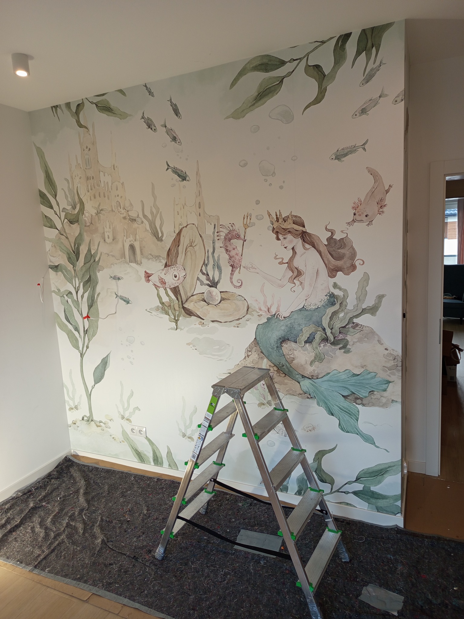 Ściana z ręcznie malowaną akwarelą: podwodny świat z syreną, zamkiem i morskimi stworzeniami. Drabina malarska stoi na podłodze, gotowa do dalszych prac.